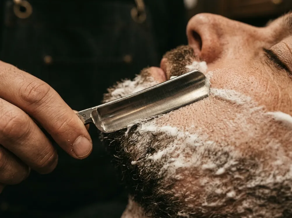 A straight razor mid-pass along a lathered jawline.